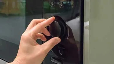 A hand turns a large black circular knob on the glossy black front panel of an appliance, with buttons visible above it.